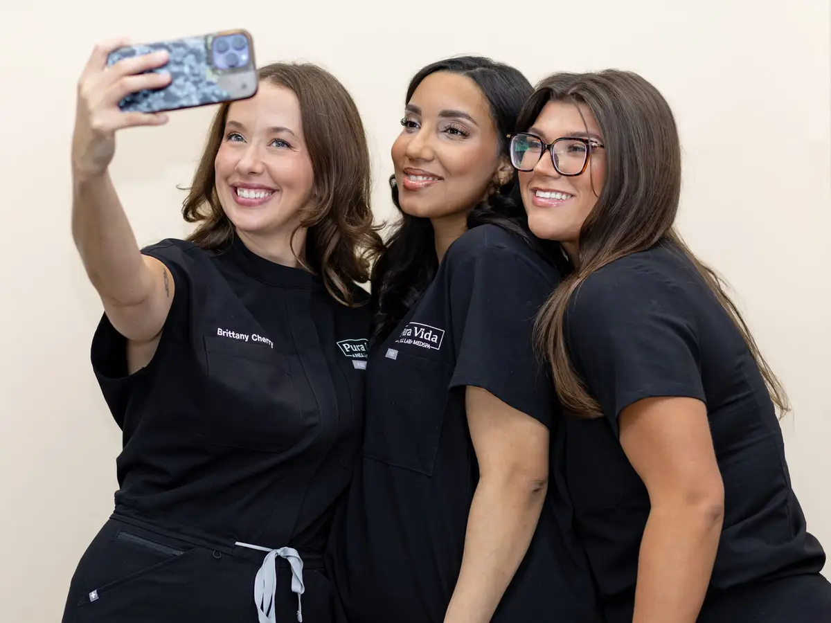 Three smiling Pura Vida team members in black scrubs taking a selfie together at the Fleming Island, FL med spa